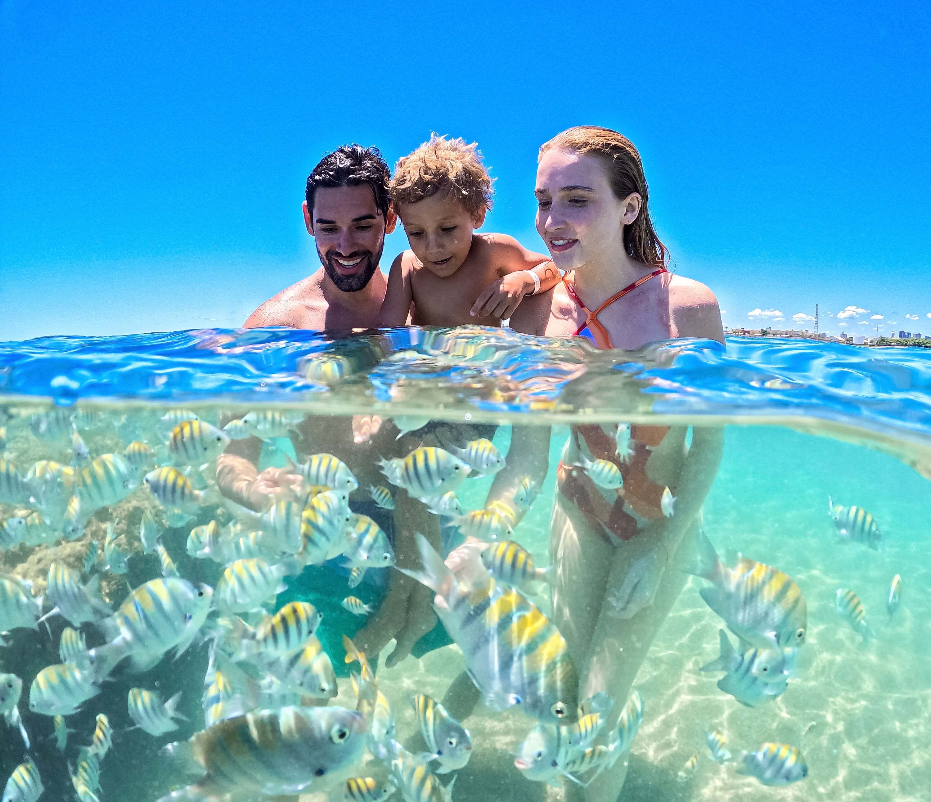 Família feliz ao lado de vários peixinhos em passeio às piscinas naturais da praia de Pajuçara, em Maceió.